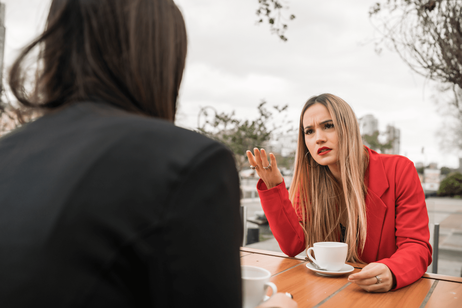 Two people talking sitting at a table outdoors over coffee, looking frustrated