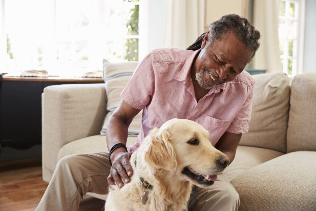 A senior person smiles and pats their happy dog while sitting on the couch.